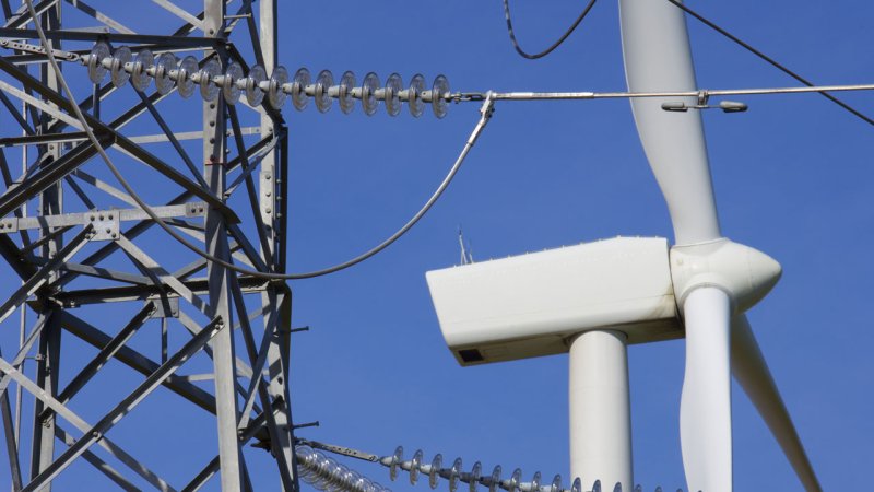 A high-voltage transmission tower with a wind turbine in the background. (Source: © pedrosala / stock.adobe.com)